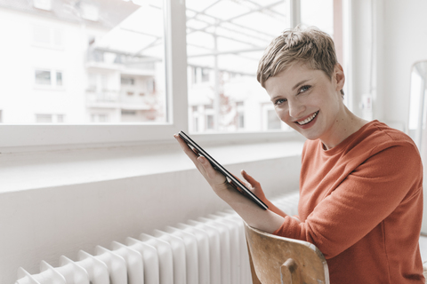 Porträt einer lächelnden Frau, die auf einem Stuhl sitzt und ein Tablet hält, lizenzfreies Stockfoto