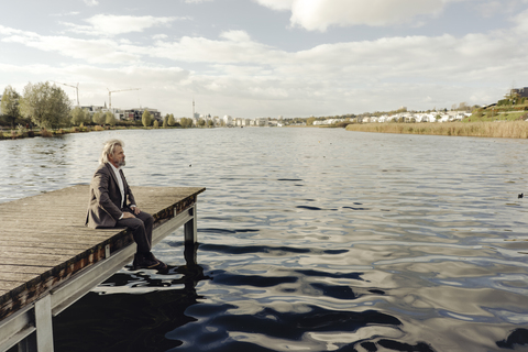 Älterer Mann sitzt auf einem Steg an einem See, lizenzfreies Stockfoto