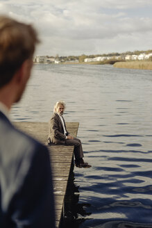 Älterer Mann sitzt auf einem Steg an einem See mit Mann im Vordergrund - KNSF03342