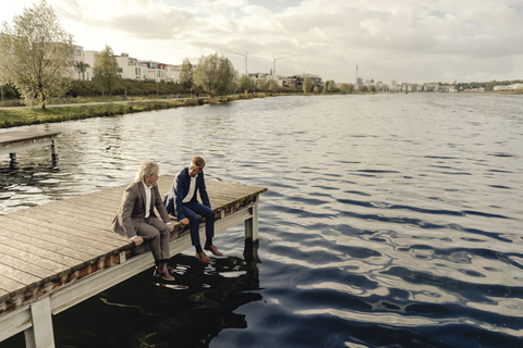 Two businessmen sitting on jetty at a lake talking stock photo