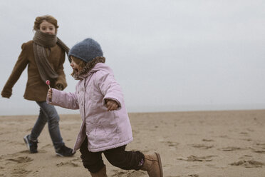 Schreiendes kleines Mädchen mit Lolli, das im Winter am Strand läuft, während ihr Bruder sie beobachtet - KMKF00096