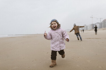 Glückliches kleines Mädchen mit Lolly, das im Winter am Strand läuft, während ihr Bruder im Hintergrund spielt - KMKF00097