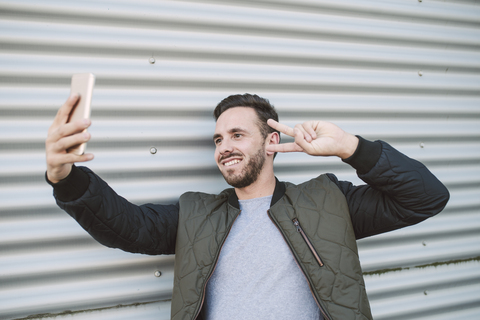 Porträt eines lächelnden Mannes, der ein Selfie mit einem Smartphone macht, das das Siegeszeichen zeigt, lizenzfreies Stockfoto