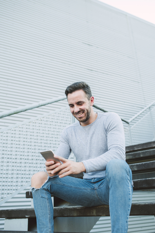 Lachender Mann sitzt auf einer Treppe und schaut auf sein Handy, lizenzfreies Stockfoto