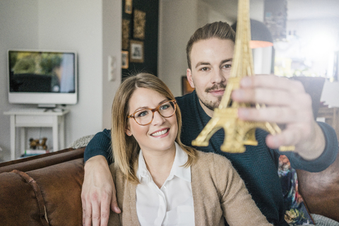 Lächelndes Paar sitzt zu Hause auf der Couch und hält ein Eiffelturm-Modell, lizenzfreies Stockfoto