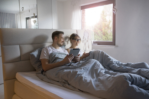 Lächelndes Paar im Bett zu Hause mit Tabletten, lizenzfreies Stockfoto