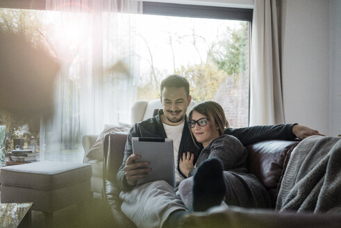 Smiling couple lying on couch at home sharing tablet - MOEF00623