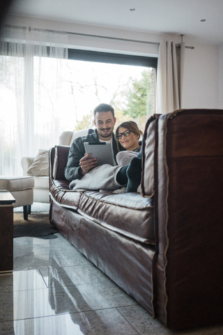 Lächelndes Paar, das zu Hause auf der Couch liegt und sich ein Tablet teilt, lizenzfreies Stockfoto