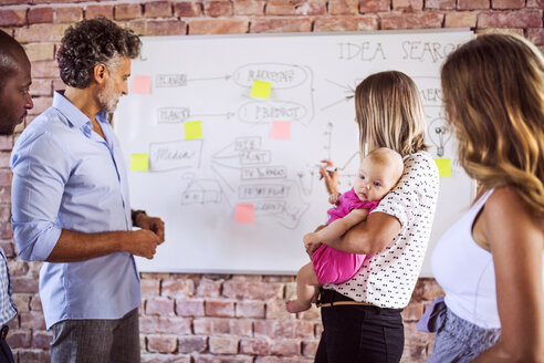 Mutter mit Baby arbeitet zusammen mit dem Team am Whiteboard an einer Backsteinmauer im Büro - HAPF02589