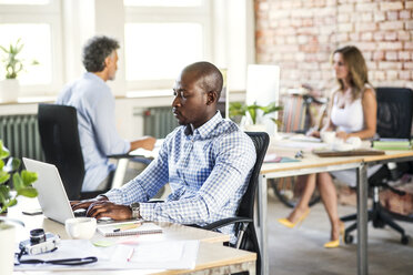 Businessman using laptop at desk in office with colleagues in background - HAPF02614