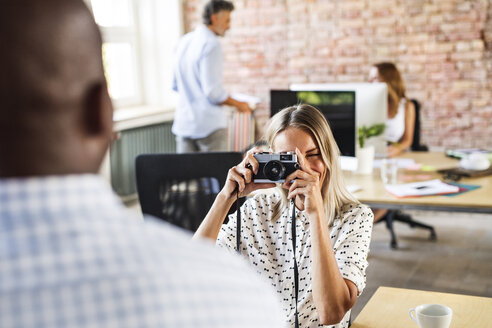 Geschäftsfrau mit Kamera, die einen Kollegen im Büro fotografiert - HAPF02616