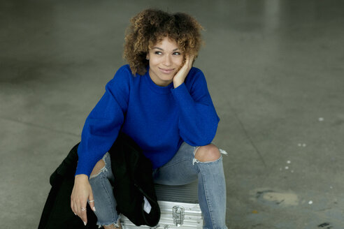 Porträt einer lächelnden Frau mit blauem Pullover, die auf einem Koffer sitzt - HHLMF00032
