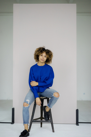 Porträt einer ernsten Frau mit blauem Pullover auf einem Hocker sitzend, lizenzfreies Stockfoto