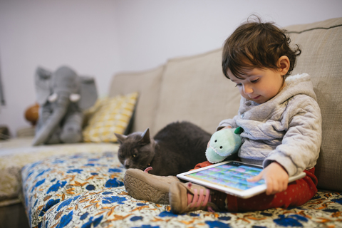 Baby-Mädchen sitzt auf der Couch und schaut Videos auf einem Tablet mit einer Katze, lizenzfreies Stockfoto