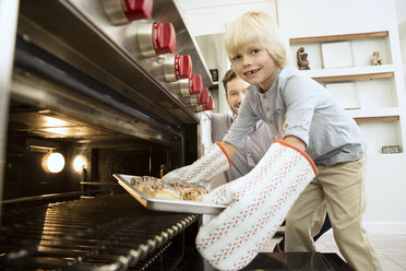 Lächelnder Junge, der ein Backblech aus dem Ofen nimmt, mit seinem Vater im Hintergrund - MFRF01074