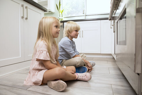 Bruder und Schwester sitzen auf dem Boden in der Küche und schauen auf den Ofen - MFRF01079