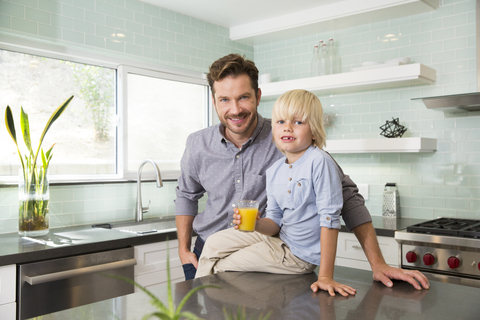 Porträt eines Jungen mit Vater in der Küche, der ein Glas Orangensaft hält, lizenzfreies Stockfoto