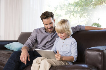 Father and son sitting on couch at home using tablet - MFRF01102