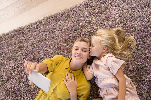 Lächelnde Mutter und Tochter liegen auf dem Teppich und machen ein Selfie, lizenzfreies Stockfoto