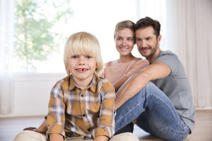Porträt eines lächelnden Jungen mit Eltern im Hintergrund, der zu Hause auf dem Boden sitzt - MFRF01124