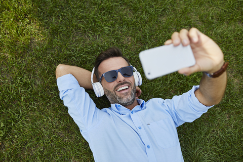 Porträt eines lachenden Mannes, der auf einer Wiese liegt und ein Selfie mit seinem Smartphone macht, Ansicht von oben, lizenzfreies Stockfoto