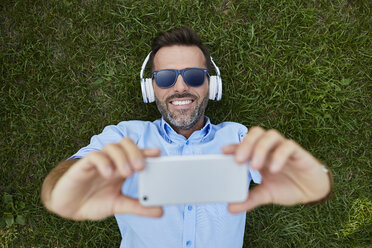 Porträt eines lachenden Mannes, der auf einer Wiese liegt und ein Selfie mit seinem Smartphone macht, Ansicht von oben - BSZF00140