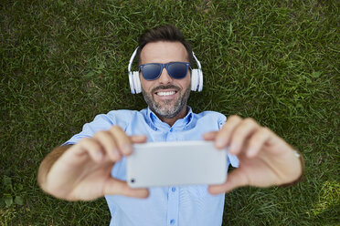 Portrait of laughing man lying on a meadow taking selfie with smartphone, top view - BSZF00140