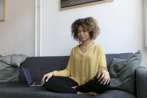Junge Frau sitzt auf der Couch zu Hause neben dem Laptop und meditiert, lizenzfreies Stockfoto