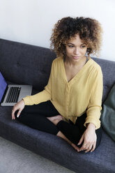 Smiling young woman sitting on couch at home next to laptop - HHLMF00059