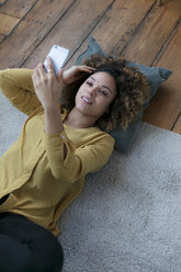 Lächelnde junge Frau auf dem Boden liegend zu Hause mit Handy - HHLMF00064