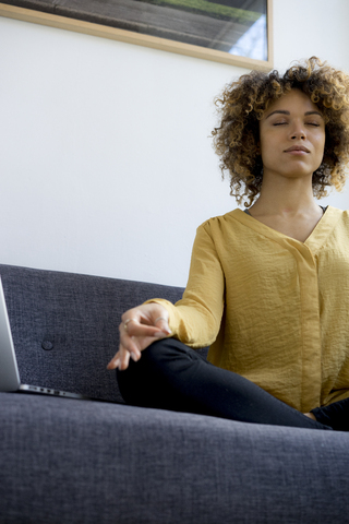 Young woman sitting on couch at home next to laptop meditating stock photo