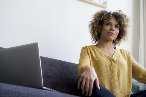 Young woman sitting on couch at home next to laptop - HHLMF00114