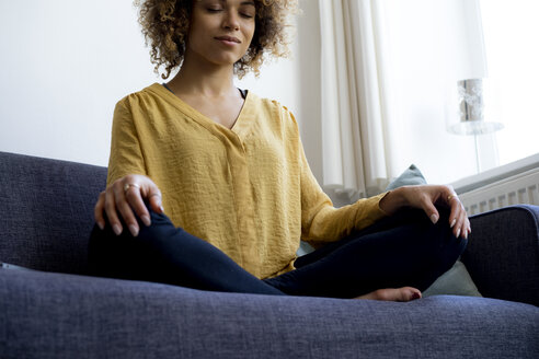 Young woman sitting on couch at home meditating - HHLMF00115