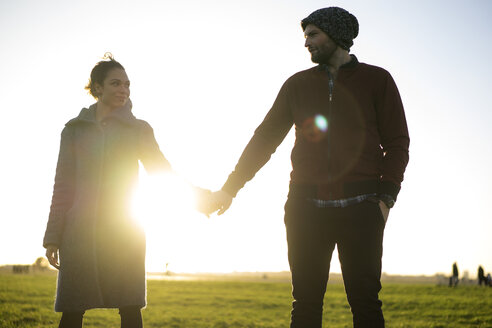 Verliebtes junges Paar, das Hand in Hand auf einer Wiese bei Sonnenuntergang steht - HHLMF00143