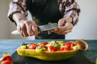 Nahaufnahme von Männerhänden, die Schokolade über eine halbe Papaya mit Banane, Kiwi und Erdbeeren schneiden - KIJF01829