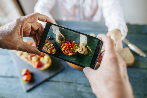 Nahaufnahme von Männerhänden, die ein Foto mit dem Mobiltelefon machen, während sie einen Salat mit Tomaten, Granatapfel, Papaya und Oliven essen, mit Papaya und Früchten an der Seite und mit einem Glas Wein - KIJF01834