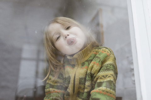 Portrait of little girl sticking out tongue while looking out of window stock photo