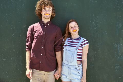 Young couple standing in front of a green wall with orange slices in their mouth stock photo