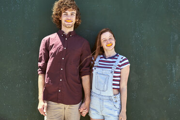 Junges Paar steht vor einer grünen Wand mit Orangenscheiben im Mund - SRYF00714
