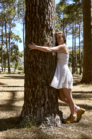 Lächelnde junge Frau umarmt einen Baum im Wald, lizenzfreies Stockfoto