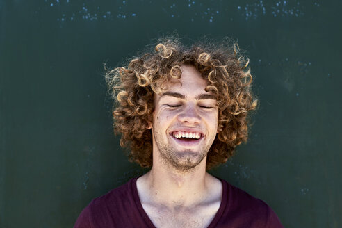 Porträt eines lachenden jungen Mannes mit lockigem Haar vor einer grünen Wand - SRYF00756