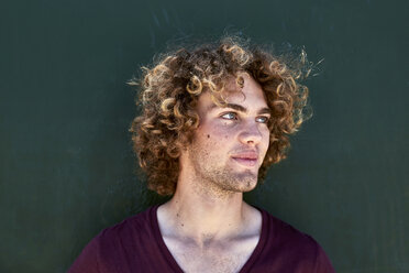 Portrait of smiling young man with curly hair in front of a green wall - SRYF00757