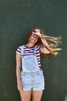 Happy young woman with windswept hair standing in front of green wall - SRYF00763