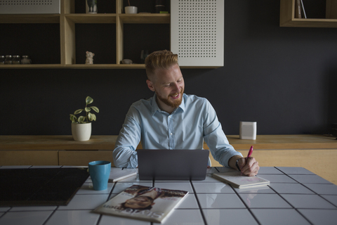 Lächelnder Geschäftsmann mit Laptop und Notizbuch bei der Arbeit zu Hause, lizenzfreies Stockfoto