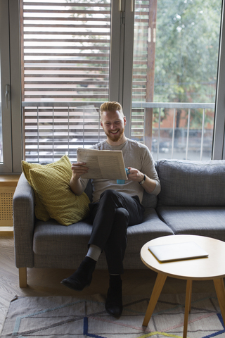 Lächelnder Mann sitzt zu Hause auf der Couch und liest Zeitung, lizenzfreies Stockfoto