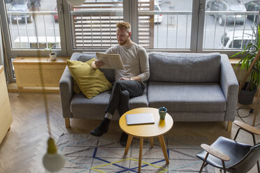 Man sitting on couch at home reading newspaper - MOMF00359