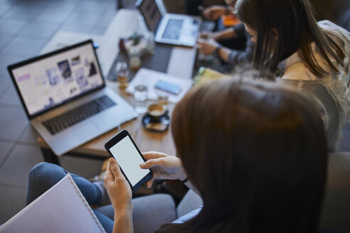 Junge Frau in einem Café, die ihr Smartphone mit Freunden benutzt, mit einem Laptop im Hintergrund - ZEDF01048