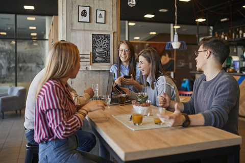 Gruppe von glücklichen Freunden, die mit Laptop, Smartphone und Getränken in einem Café zusammensitzen, lizenzfreies Stockfoto