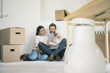 Couple sitting in new home surrounded by cardboard boxes looking at tablet - MOEF00681