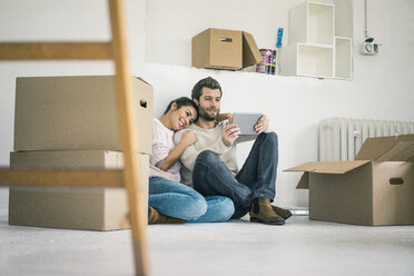 Couple sitting in new home surrounded by cardboard boxes looking at tablet - MOEF00682
